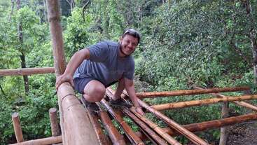 Un homme souriant, vêtu d'un t-shirt gris et d'un short, construit une structure en bambou dans une forêt luxuriante et verdoyante. Il semble heureux et enthousiaste à l'idée de ce projet.