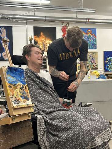 Un homme en fauteuil roulant, recouvert d'une couverture à motifs, peint avec sa bouche tandis qu'un autre homme l'assiste. Des œuvres d'art colorées décorent le fond de l'atelier.