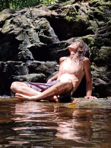 Un homme aux cheveux longs est assis sur un rocher en eau peu profonde, torse nu, la tête penchée en arrière et les yeux fermés, se prélassant au soleil.