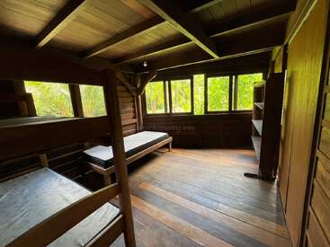 L'image représente une chambre rustique avec des murs et un plafond en bois, comprenant deux lits superposés, un mur avec fenêtre et un plancher en bois, entourée d'une végétation luxuriante à l'extérieur.