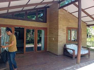 L'image représente un homme debout sur une terrasse en bois à l'extérieur d'une maison en briques, avec un canapé et de grandes fenêtres, entourée d'une végétation luxuriante.