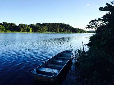 L'image représente une scène sereine d'un petit bateau flottant sur un plan d'eau tranquille, entouré d'une végétation luxuriante et d'un ciel bleu clair.