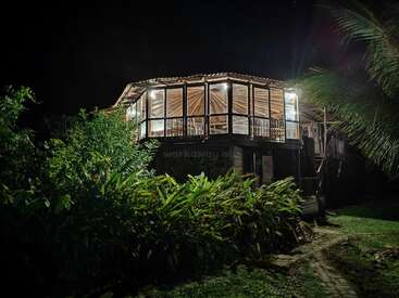 L'image représente une maison à deux étages avec un balcon, entourée d'une végétation luxuriante et de palmiers, sur fond de ciel nocturne sombre.