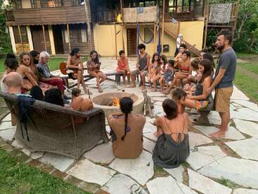 Un groupe de personnes, dont des enfants, se rassemblent autour d'un foyer dans une arrière-cour, certaines assises sur des chaises, d'autres à même le sol.