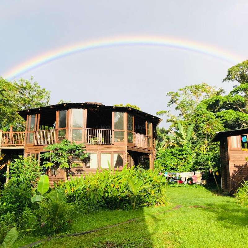L'image représente une maison en bois sereine avec un porche enveloppant, entourée d'une verdure luxuriante et d'un arc-en-ciel vibrant au-dessus d'elle, sur fond de ciel gris.