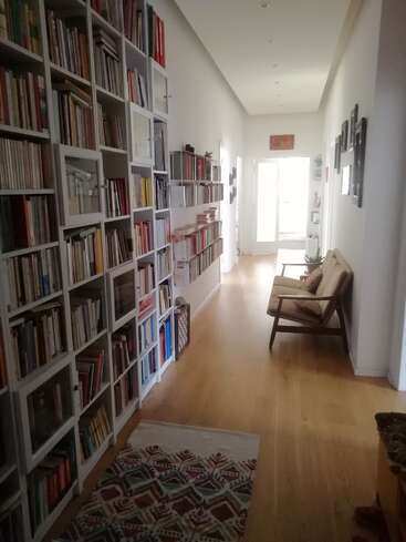 Ein heller Flur mit Holzfußboden, der mit mehreren Bücherregalen voller Bücher ausgestattet ist. Es gibt einen gemütlichen Stuhl, einen gemusterten Teppich, gerahmte Bilder und ein großes Fenster.