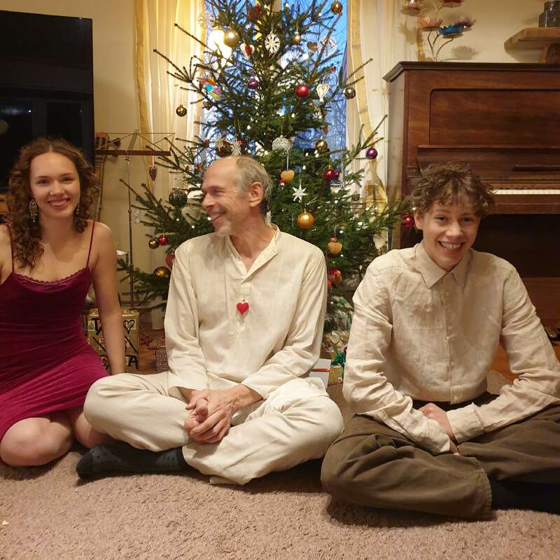 Tres personas están sentadas felices delante de un árbol de Navidad decorado. Sonríen, van bien vestidas y hay regalos y un piano al fondo. Se respira un ambiente festivo y acogedor.