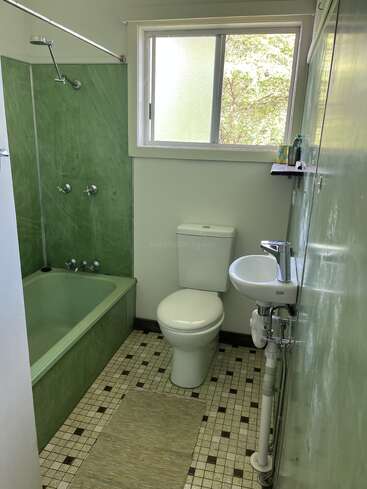 Cette salle de bains comprend des carreaux verts, une baignoire avec douche, des toilettes, un petit lavabo mural, une fenêtre, une étagère avec des articles de toilette et un sol en carreaux à motifs.