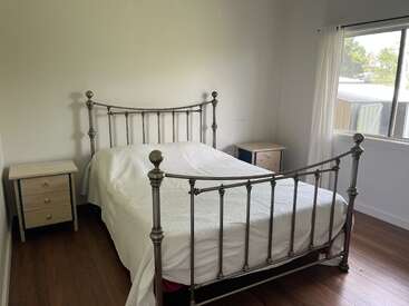 Cette chambre comprend un cadre de lit en métal avec un couvre-lit blanc, deux tables de nuit en bois, des planchers en bois, des murs blancs et une grande fenêtre avec des rideaux transparents.