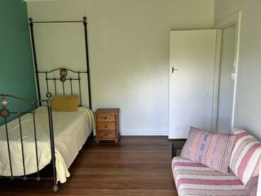 Cette image montre une chambre simple avec un lit simple à armature métallique, une table de nuit en bois, une porte blanche ouverte et un canapé à rayures sur un sol en bois.