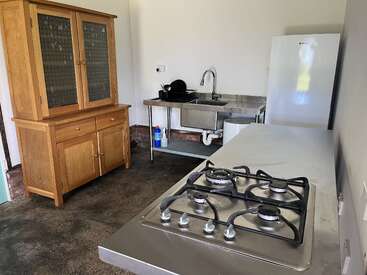 Cette image montre une cuisine simple avec un meuble en bois, un plan de travail et un évier en acier inoxydable, une cuisinière à gaz, un réfrigérateur et un égouttoir à vaisselle. Le sol est en béton.