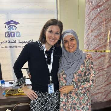 Two women smiling, standing side by side indoors. One wears a hijab and patterned dress; the other, a lanyard and black shirt. Friendly, warm atmosphere, educational setting.