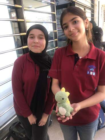 Two smiling girls wearing matching maroon uniforms stand together. One holds a small, handmade green crocheted dinosaur toy. They appear happy, posing outside a building.