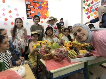 A group of smiling children and their teacher pose in a colorful classroom decorated with paper flowers. Tables are filled with various fruits and sunflower arrangements.