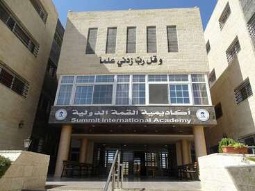 The image shows the entrance of Summit International Academy, a beige stone building with both Arabic and English signage, large windows, and a welcoming stairway.