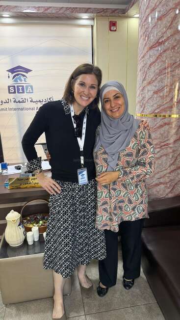 Two women stand smiling together inside an office. One wears a black dress, the other a patterned tunic and hijab. Behind them is a Summit International Academy banner.