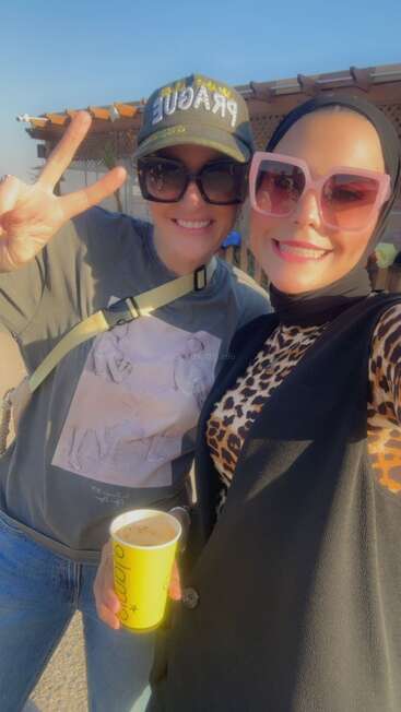 Two women smile for a cheerful selfie outdoors. Both wear stylish sunglasses. One holds a coffee cup, the other gestures a peace sign, enjoying sunny weather.