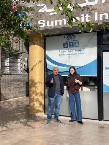 Zwei Menschen stehen lächelnd vor der Summit International Academy und zeigen die Daumen nach oben. Ein sonniger Tag, Pflanzen mit rosa Blüten umrahmen die Szene und schaffen eine einladende Atmosphäre.