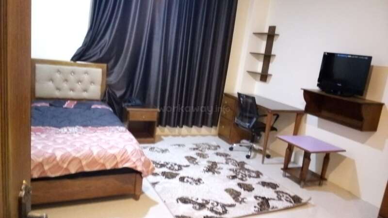 This room features a single bed with a tufted headboard, dark curtains, a desk with a chair, a TV, shelves, a nightstand, and a patterned rug.