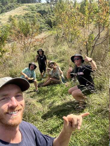 Five smiling people pose outdoors on grassy land among young trees, enjoying sunny weather. They all wear hats and look happy, making playful gestures towards the camera.