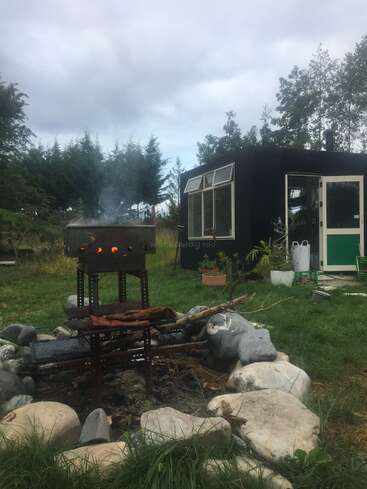A rustic outdoor scene shows a black tiny house, a fire pit surrounded by rocks, and a metal grill with logs, nestled in green countryside.