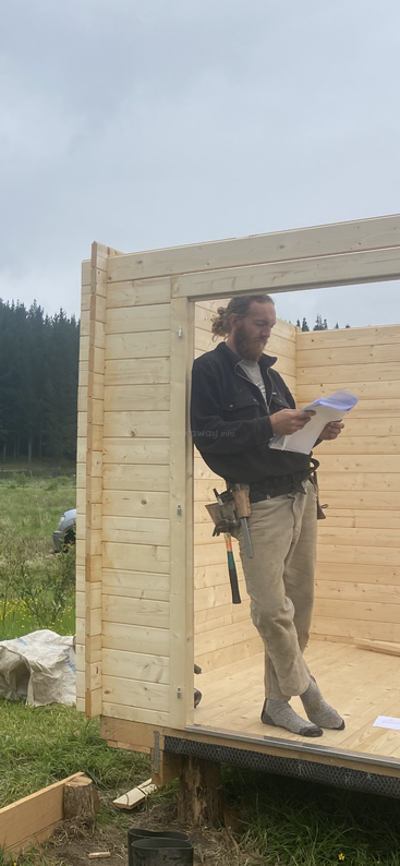 A man stands inside a partially built wooden cabin, reading papers. He has a tool belt, wears socks, and construction is ongoing in a rural outdoor setting.