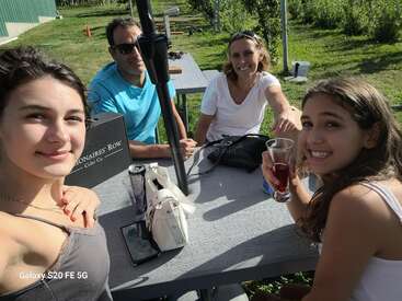 Una familia de cuatro miembros disfruta de un día soleado al aire libre, sentados en una mesa de picnic con bebidas, sonriendo y posando juntos para un alegre selfie.
