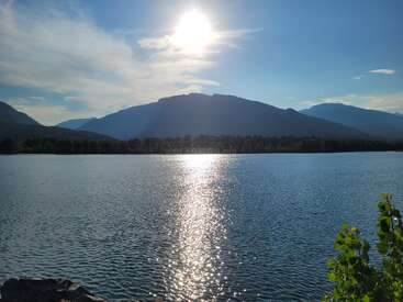 The image shows a serene lake reflecting bright sunlight, surrounded by mountains, forests, and clear blue sky, with some greenery in the foreground. Peaceful and beautiful.