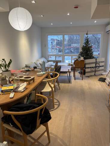 Ein heller Wohn-Essbereich mit Holzböden, einem langen Tisch, ein paar Sachen rumstehend, modernen Stühlen, einer Papierlaterne, einem Sofa, einem Weihnachtsbaum, großen Fenstern und Blick nach draußen.