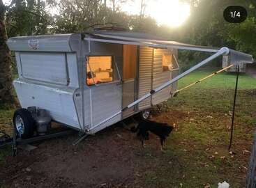 A vintage silver caravan with an awning sits in a grassy, wooded area at sunset. Warm lights glow inside. A black dog stands beneath the awning.