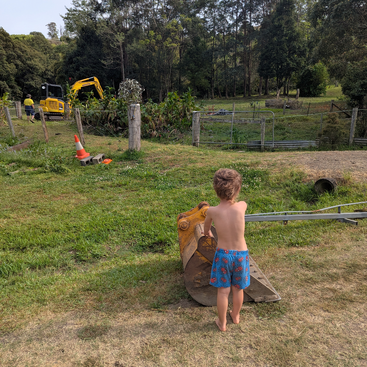 A young boy in blue shorts stands barefoot, touching a large excavator bucket. In the background, workers and a yellow excavator are visible near green trees.