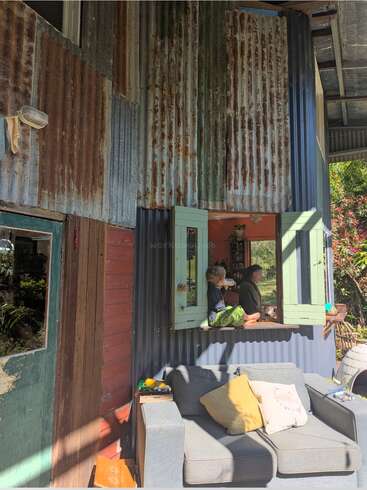 Rustic house exterior with corrugated metal walls, open green window, two people inside, gray outdoor couch with yellow and white cushions, sunlight, and surrounding greenery.