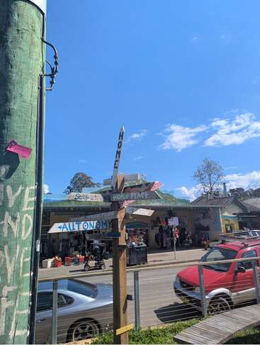 A sunny street scene with shops, cars, and a wooden signpost featuring words like “HOME,” “WORLD PEACE,” and “AUTONOMY.” Blue sky and scattered clouds overhead.