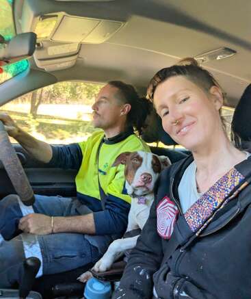A man is driving a car, a woman is in the passenger seat smiling at the camera, and a brown and white dog is in between them.