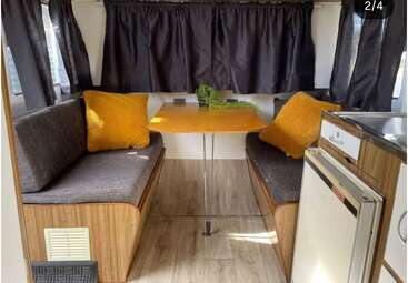 This cozy camper interior features two cushioned benches, a wooden table, yellow pillows, dark curtains, wood paneling, a small refrigerator, and a plant centerpiece.
