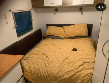 A cozy caravan bedroom with a neatly made bed covered in yellow bedding, two pillows, overhead cabinets, a window, small table, and soft lighting.