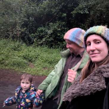 A family of three walks outdoors. The woman smiles at the camera, all wearing warm clothes and hats. Greenery surrounds them, creating a cozy, natural setting.