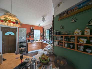 This cozy kitchen features wooden cabinets, a green accent wall with shelves of jars, a large window, hanging lamp, and various kitchen items on the counter.