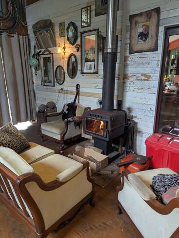 A cozy living room with vintage armchairs, a wood stove burning, mirrors and art on white wooden walls, a guitar, and various eclectic decorations scattered around.