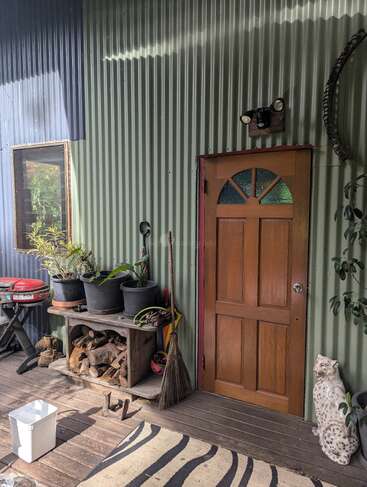 A rustic porch with a wooden door, potted plants, broom, wood storage, animal statue, outdoor furniture, a zebra-striped rug, and corrugated metal wall, sunlit and cozy.