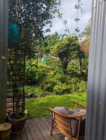A cozy outdoor deck with a wicker chair overlooks a lush, green garden filled with plants and trees, inviting peace and relaxation in nature.