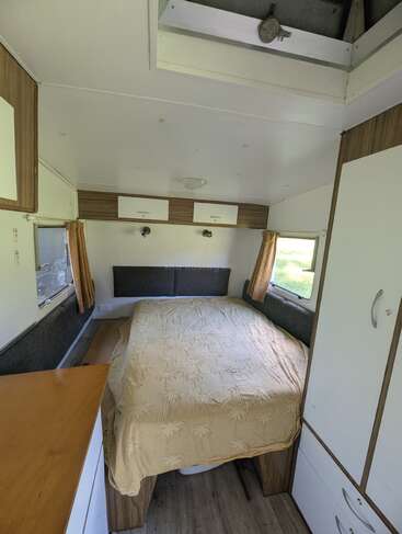 This cozy camper bedroom features a double bed with a beige blanket, wood and white storage cabinets, small windows with curtains, and a skylight overhead.