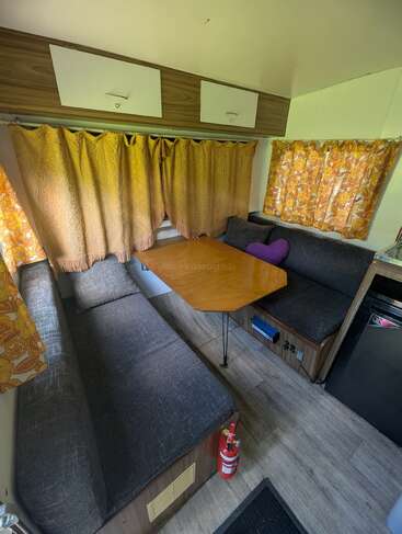 This image shows a cozy camper interior with gray cushioned benches, a wooden table, patterned yellow curtains, overhead cabinets, a fridge, and a fire extinguisher on the floor.