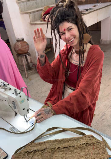 Eine lächelnde Frau mit Dreadlocks, Gesichtsbemalung und Tattoos winkt, während sie an einer Nähmaschine sitzt und in einem kreativen Arbeitsbereich einen roten Bademantel trägt.