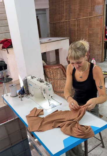 Eine Frau sitzt an einer Nähmaschine und arbeitet in einem Arbeitsraum mit braunem Stoff. Sie wirkt konzentriert, umgeben von Werkzeugen, Stühlen und einer Treppe im Hintergrund.