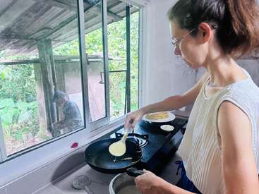 Une femme prépare des crêpes sur la cuisinière, en versant la pâte dans une poêle. Devant la fenêtre, un homme est assis et travaille, entouré de verdure.