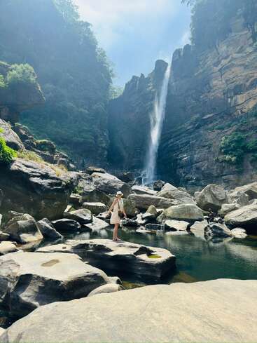 Une femme se tient debout sur un gros rocher, entourée d'eau et de rochers, tandis qu'une haute cascade dévale un canyon escarpé et luxuriant sous un ciel radieux.