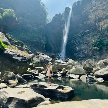 A woman stands on a large rock surrounded by water and boulders, with a tall waterfall cascading down a steep, lush canyon under a bright sky.