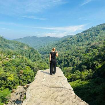 A person stands on a rocky ledge overlooking a lush, green forest valley under a bright blue sky with scattered clouds, surrounded by distant mountains.