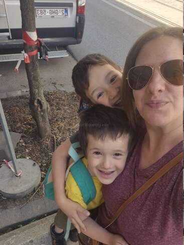 Una mujer con gafas de sol abraza a dos niños sonrientes, uno de ellos con una mochila. Parecen felices, posando para un selfie en una acera de la ciudad cerca de una furgoneta.